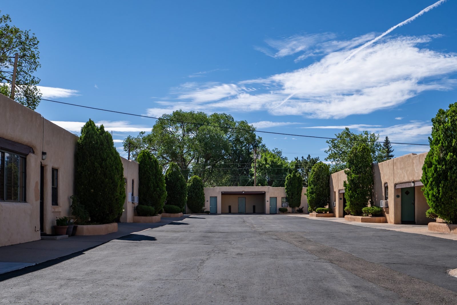 Looking down the U-shaped courtyard with adobe walls, green doors, and tall trees under a clear New Mexico sky