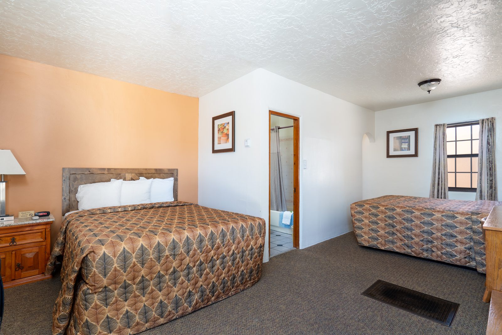 Two-queen guest room with two made beds, bathroom doorway, and a window with curtains